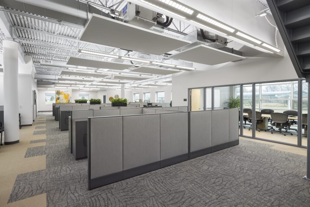 Modern office interior featuring rows of grey fabric cubicles, suspended acoustic ceiling panels, and a glass-walled conference room