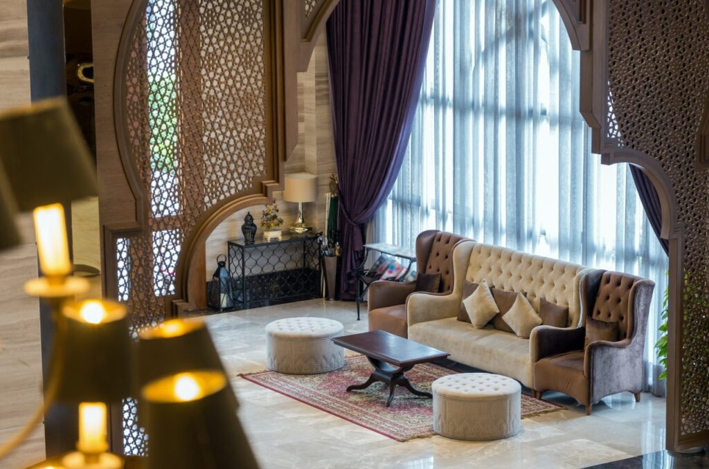 Lounge area with a tufted sofa, two round ottomans, and a coffee table, framed by decorative wooden arches and floor-to-ceiling curtains