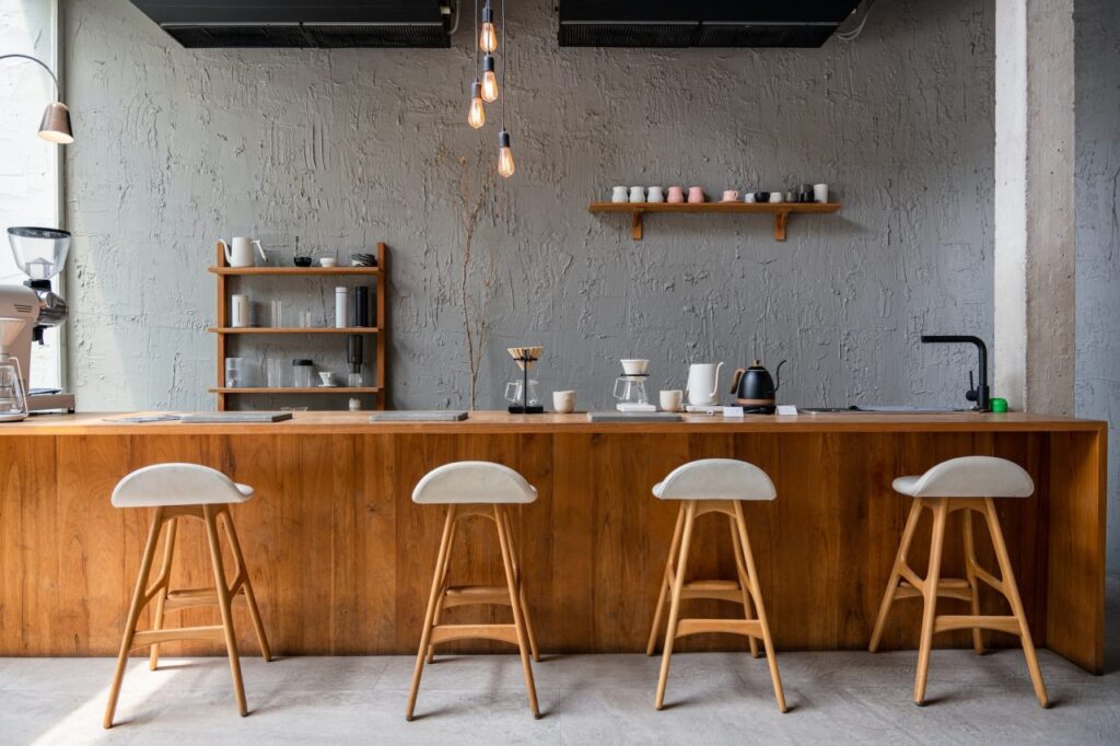 Modern coffee bar with a wooden counter, four white stools, and shelves displaying brewing equipment against a textured gray wall