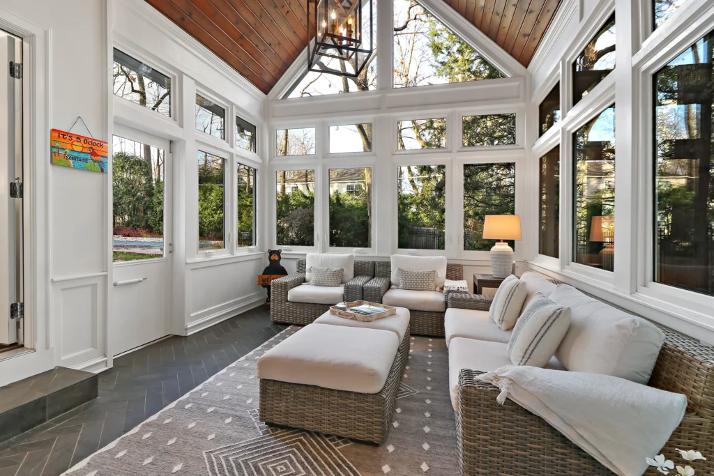 Sunroom with white walls, a vaulted wood ceiling, and floor-to-ceiling windows, featuring wicker furniture with white cushions and a patterned rug