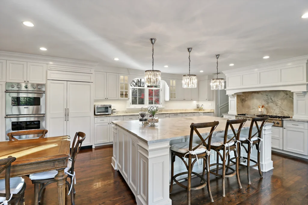 Spacious white kitchen with a large marble-topped island, three crystal pendant lights, and dark wood flooring
