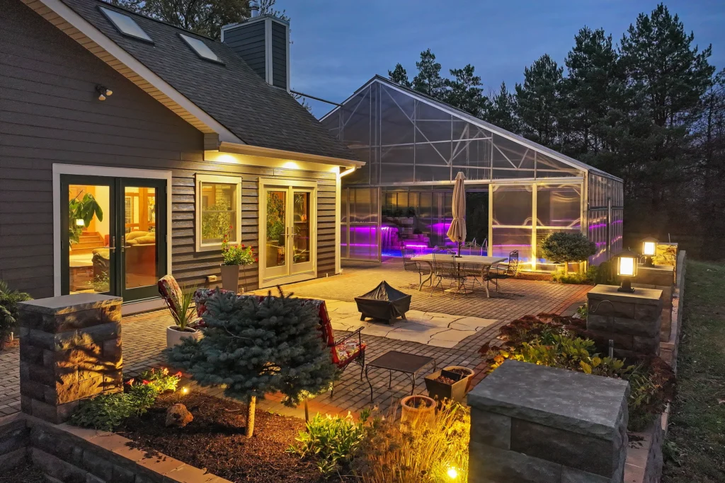 A backyard patio at dusk featuring a brick seating area with a fire pit, outdoor furniture, and a large attached greenhouse containing a glowing pool