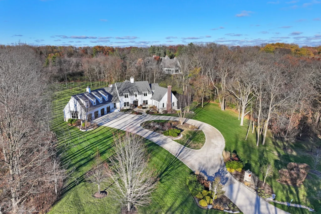 Aerial view of a white multi-gabled house with a circular driveway, surrounded by a large lawn and a wooded area in autumn