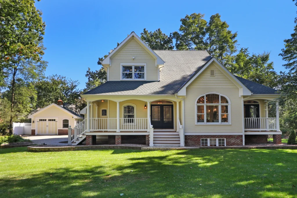 Yellow two-story house with a wrap-around porch, dark trim, and a detached garage on a sunny day