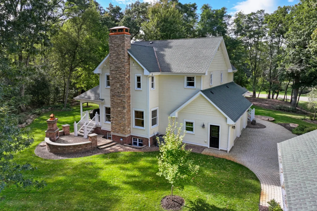 Aerial view of a two-story yellow house with a stone chimney, green roof, and a stone patio with an outdoor fireplace