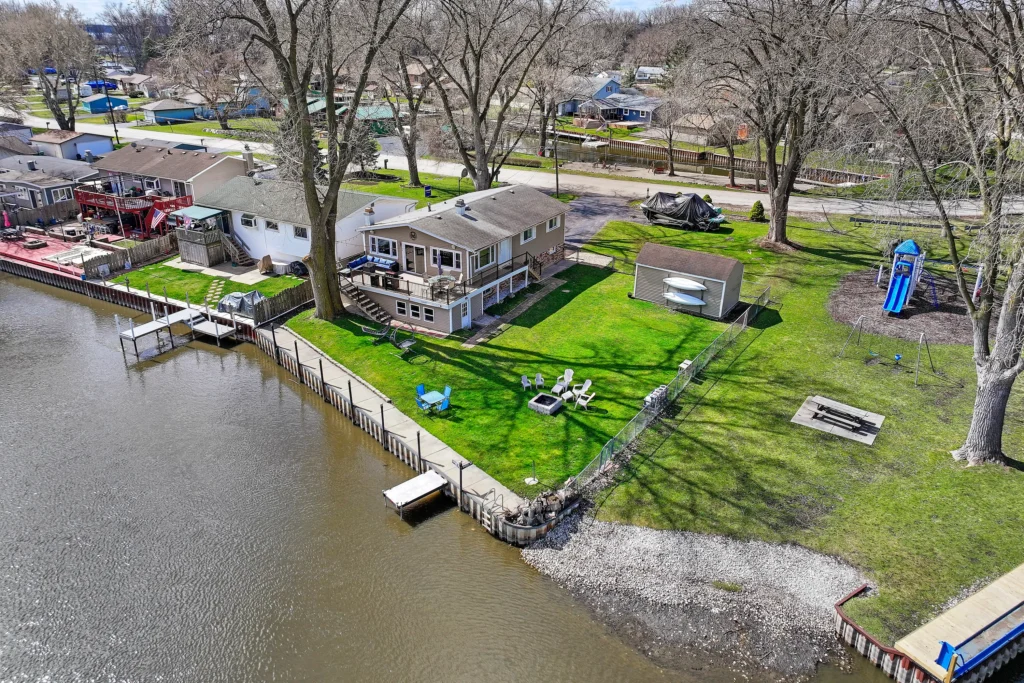 Aerial view of a waterfront house with a large backyard, deck, fire pit, and private dock along a canal