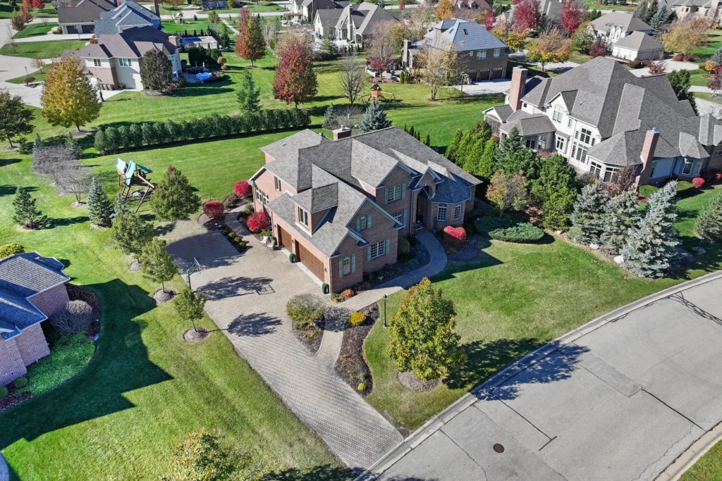 Aerial view of a suburban neighborhood featuring large brick homes with spacious lawns, paved driveways, and mature trees during autumn