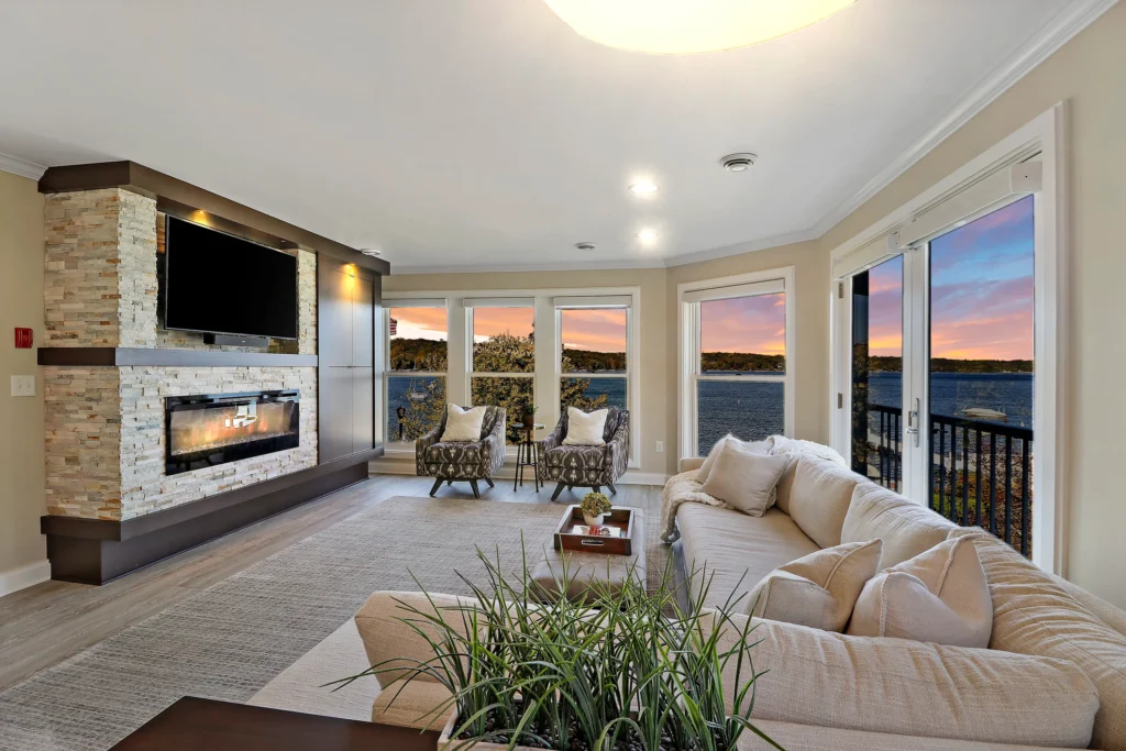 Living room with a stone fireplace, large sectional sofa, and floor-to-ceiling windows overlooking a lake at sunset