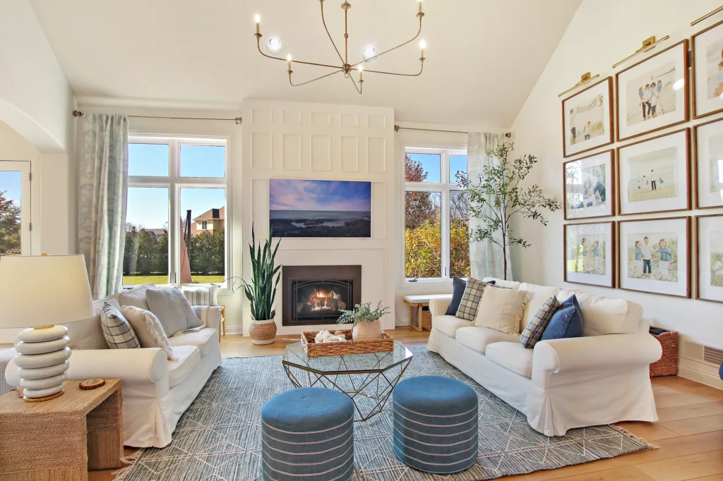 Bright living room with two white sofas, a central fireplace, a geometric coffee table, and a gallery wall of family photos