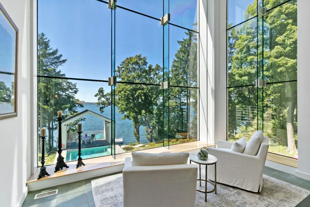 Two white armchairs face a floor-to-ceiling window overlooking a lake, a small pool, and a covered patio